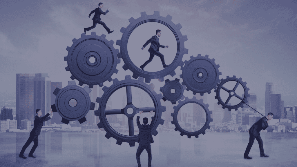 Five sharply dressed men with short brown hair are seen working with giant gears in an urban setting during the day. They are showing engineering productivity and engineering capability. One pushes a gear, another pulls a gear with a rope, one balances a gear on their back, one is running inside the gear, and another is running on top of the gear. This image has a sophisticated mood and could be used in projects about teamwork, business, and corporate operations.