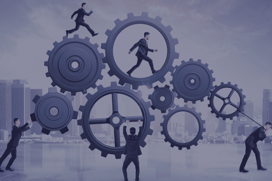Five sharply dressed men with short brown hair are seen working with giant gears in an urban setting during the day. They are showing engineering productivity and engineering capability. One pushes a gear, another pulls a gear with a rope, one balances a gear on their back, one is running inside the gear, and another is running on top of the gear. This image has a sophisticated mood and could be used in projects about teamwork, business, and corporate operations.