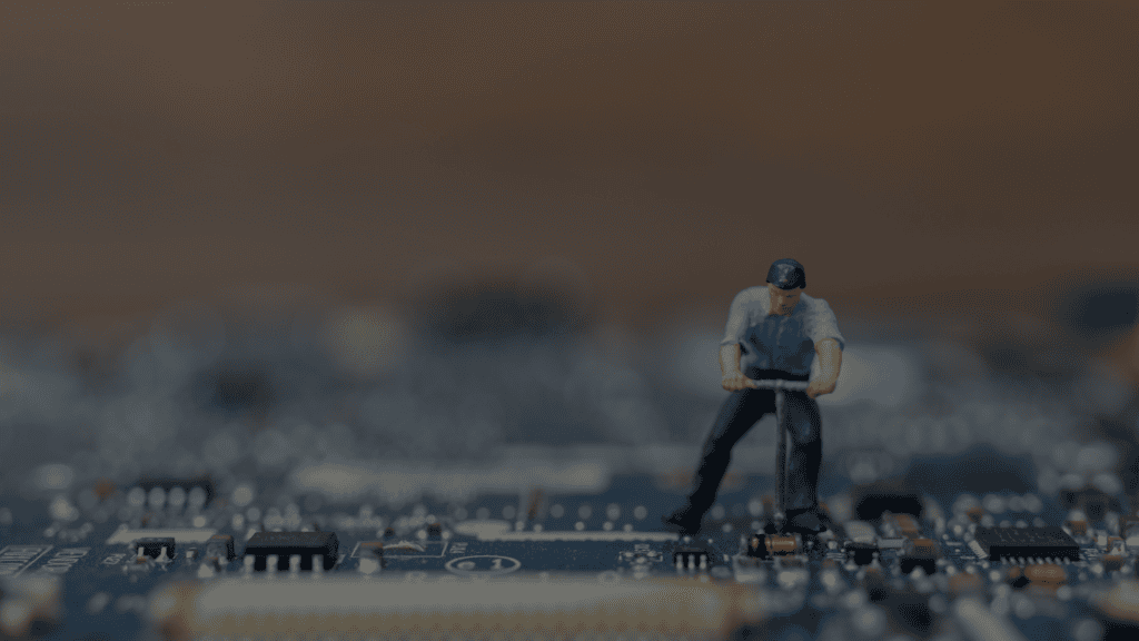 A tiny figurine of a man with short brown hair, wearing a blue short-sleeved shirt, dark trousers and a blue baseball cap, uses a tool on a blue circuit board. He is a product engineer. The image evokes the idea of fixing or inspecting computer or technology components. The background is out of focus. The mood is serious and analytical. This photo could be used for illustrative purposes in business or educational materials about technology, engineering, or innovation.
