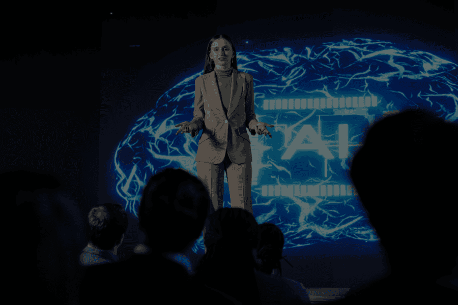 An adult woman with auburn hair stands on a stage in front of a glowing blue graphic, speaking to an audience. She wears a stylish tan suit and a green turtleneck. The glowing graphic behind her has the letters 'AI' prominently displayed. The audience sits in the dark, illuminated by the stage lights. This photo could be used for projects relating to technology, artificial intelligence, corporate settings, and public speaking. She is talking about AI strategy, pilots, AI ROI and product engineering.